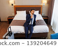 Tired Businessman Sitting on Hotel Bed After Business Trip with Suitcase in Modern Room 139204649