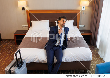 Tired Businessman Sitting on Hotel Bed After Business Trip with Suitcase in Modern Room Tired Businessman Sitting on Hotel Bed After Business Trip with Suitcase in Modern Room 139204649