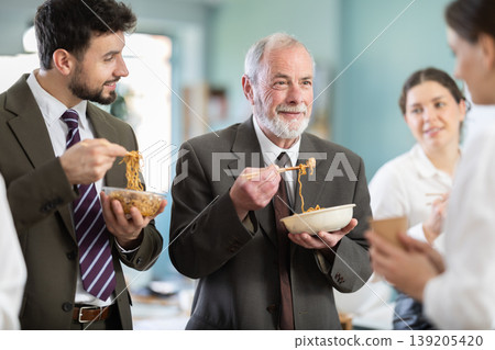 Group of colleagues, man and woman, are having lunch in office during their break, eating noodles Group of colleagues, man and woman, are having lunch in office during their break, eating noodles 139205420