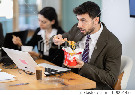 Young businessman having takeout meal during lunch break at work sitting at workplace at laptop with other colleagues in office Young businessman having takeout meal during lunch break at work sitting at workplace at laptop with other colleagues in office 139205453