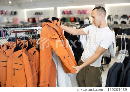Guy stopped near showcase with outerwear, holds jacket hanger in hands and examines product Guy stopped near showcase with outerwear, holds jacket hanger in hands and examines product 139205489