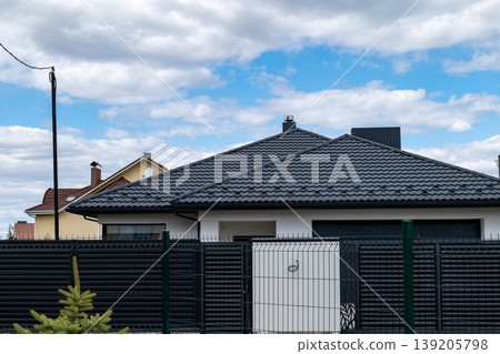 A modern house with a black roof stands within a fenced area on a cloudy day in a suburban neighborhood A modern house with a black roof stands within a fenced area on a cloudy day in a suburban neighborhood 139205798