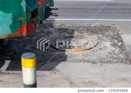 Road works are taking place on a city street. A construction vehicle is parked next to a manhole cover. The area shows signs of work Road works are taking place on a city street. A construction vehicle is parked next to a manhole cover. The area shows signs of work 139205806