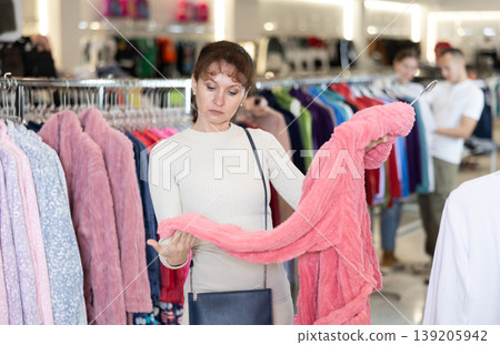 Woman stopped near leisure wear showcase, holds eiderdown bathrobe hanger in hands, examines product Woman stopped near leisure wear showcase, holds eiderdown bathrobe hanger in hands, examines product 139205942