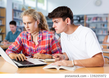 Positive female and male students teenagers study together at laptop while sitting in school library Positive female and male students teenagers study together at laptop while sitting in school library 139205972