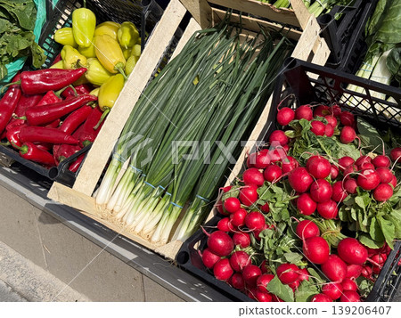 Fresh radishes, scallions, peppers and leafy greens displayed in crates outside a shop. Vegetable retail, seasonal produce, grocery shopping and local food market freshness. Fresh radishes, scallions, peppers and leafy greens displayed in crates outside a shop. Vegetable retail, seasonal produce, grocery shopping and local food market freshness. 139206407