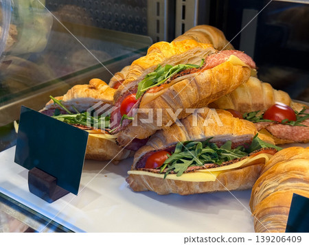 Croissant sandwiches with cheese, salami, and arugula in display case. Bakery assortment, ready food choice, snack purchase, and cafe offering with layered ingredients. 139206409