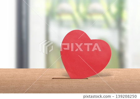 A red heart symbol being inserted into a slot on a wooden surface, representing charity, donation, kindness, support, and organ donation concepts in a blurred indoor setting A red heart symbol being inserted into a slot on a wooden surface, representing charity, donation, kindness, support, and organ donation concepts in a blurred indoor setting 139208250