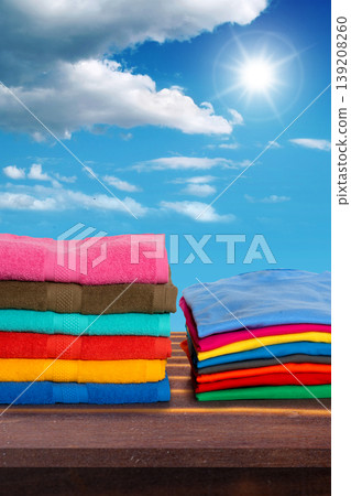 Two neat stacks of brightly colored laundry, including towels and t-shirts, sit on a wooden shelf against a backdrop of a clear blue sky with white clouds and a bright shining sun Two neat stacks of brightly colored laundry, including towels and t-shirts, sit on a wooden shelf against a backdrop of a clear blue sky with white clouds and a bright shining sun 139208260
