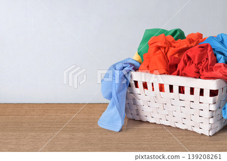 A white woven laundry basket overflowing with brightly colored clothes like red, blue, green, and orange, sitting on a wooden tabletop against a clean light background 139208261