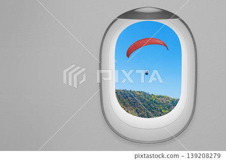 A view of a red paraglider soaring over forested mountains as seen through the oval window of an airplane against a clear blue sky, illustrating travel and adventure A view of a red paraglider soaring over forested mountains as seen through the oval window of an airplane against a clear blue sky, illustrating travel and adventure 139208279