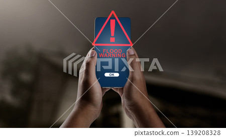 A hand holds a smartphone showing a flood warning alert, with an overturned car partially submerged in water under a dark, stormy sky in the background 139208328