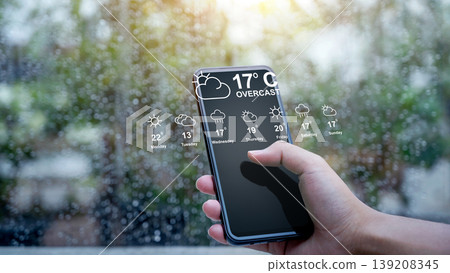Close-up of a hand holding a smartphone showing a weather forecast app interface with temperatures and icons, set against a blurry rainy day background through a window Close-up of a hand holding a smartphone showing a weather forecast app interface with temperatures and icons, set against a blurry rainy day background through a window 139208345