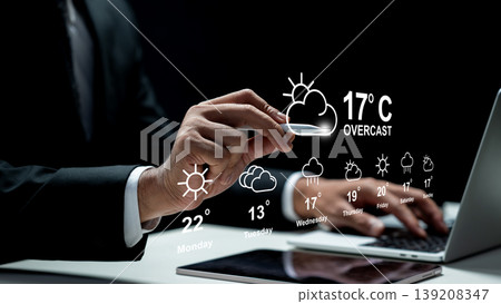A professional in a suit interacts with a digital holographic weather forecast display showing temperatures and icons over a laptop and tablet on a desk A professional in a suit interacts with a digital holographic weather forecast display showing temperatures and icons over a laptop and tablet on a desk 139208347