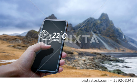 A hand holds a smartphone showing a cloudy weather app forecast with 22C against the dramatic, rugged mountain peaks and coastline of Vestrahorn, Iceland A hand holds a smartphone showing a cloudy weather app forecast with 22C against the dramatic, rugged mountain peaks and coastline of Vestrahorn, Iceland 139208348