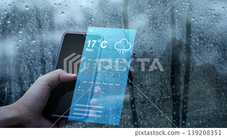 A hand holds a smartphone displaying a futuristic holographic augmented reality weather forecast interface with rain icons and temperature, set against a wet window on a rainy day 139208351