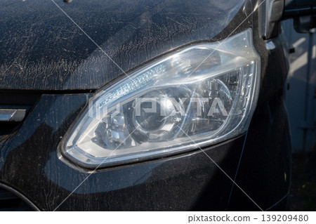 A black vehicle with a dirty exterior is parked in a car lot. The focus is on the headlight that is not clean A black vehicle with a dirty exterior is parked in a car lot. The focus is on the headlight that is not clean 139209480