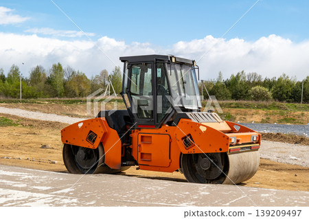 A roller is resting on a dirt road at a construction site near green trees on a sunny day with some clouds in the sky A roller is resting on a dirt road at a construction site near green trees on a sunny day with some clouds in the sky 139209497