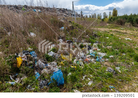 Litter is present on the ground among grass and bushes with a cloudy sky above and a distant landfill area visible Litter is present on the ground among grass and bushes with a cloudy sky above and a distant landfill area visible 139209501