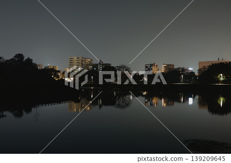 Under a dark, atmospheric sky, the quiet lake reflects the faint light of low-rise buildings at night. Under a dark, atmospheric sky, the quiet lake reflects the faint light of low-rise buildings at night. 139209645