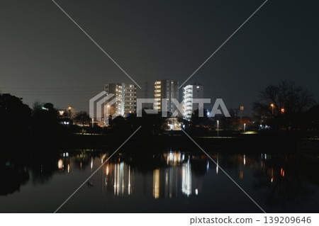 Under a dark, atmospheric sky, the quiet lake reflects the faint light of low-rise buildings at night. Under a dark, atmospheric sky, the quiet lake reflects the faint light of low-rise buildings at night. 139209646