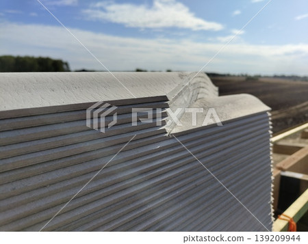 Stack of corrugated fiber cement roofing sheets with layered edges and dust texture on outdoor construction site under blue sky. Roofing, construction, building materials, exterior installation 139209944