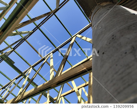 Wooden roof frame structure with beams and trusses viewed from below against clear blue sky during construction. Construction, carpentry, architecture, engineering, housing development Wooden roof frame structure with beams and trusses viewed from below against clear blue sky during construction. Construction, carpentry, architecture, engineering, housing development 139209956