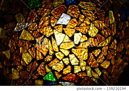 Close-up of colorful mosaic glass texture with warm light, glowing amber and gold stained glass fragments for decorative artistic background. Close-up of colorful mosaic glass texture with warm light, glowing amber and gold stained glass fragments for decorative artistic background. 139210194