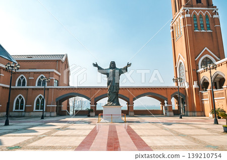 Jesus Christ statue with outstretched arms stands in a sunny plaza before a brick church, overlooking water and a distant city 139210754