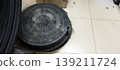 Dirty black composite manhole cover lies on a white tiled floor. The cover has distinctive mesh pattern, stylized "I" in the center, numerous bolt holes around the perimeter 139211724
