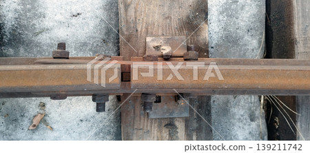 Top-down view shows a rusty railway track joint secured with heavy iron bolts and plates. The rail rests on a weathered wooden sleeper, surrounded by dirty, melting snow during the spring thaw 139211742