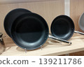 Three professional black non-stick frying pans with polished chrome handles are displayed on a wooden shelf. The sleek kitchenware is organized neatly, showcasing different sizes for culinary use 139211786