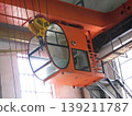 Inside an industrial building, an orange overhead crane cabin is suspended from massive steel beam. Curved glass windows provide the operator with excellent visibility, revealing thick cables 139211787