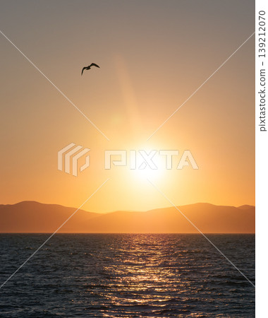 Sun dips below distant mountains painting the vast ocean with a warm, fiery glow at dusk Sun dips below distant mountains painting the vast ocean with a warm, fiery glow at dusk 139212070