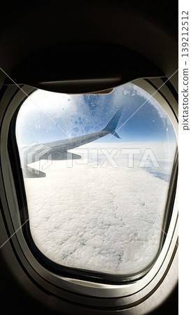 Airplane wing view from plane window over blue sky landscape in sunlight. Airplane porthole. Summer travel background. Vertical Airplane wing view from plane window over blue sky landscape in sunlight. Airplane porthole. Summer travel background. Vertical 139212512