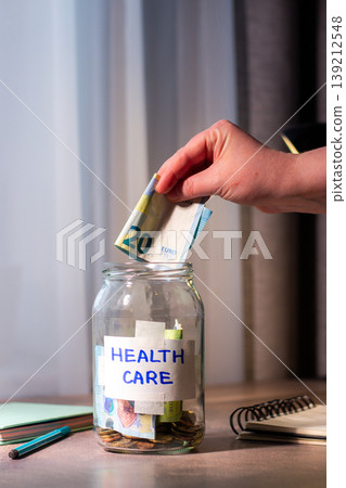 Hand dropping money into glass savings jar on table. Close up of fingers inserting cash money into transparent container for Health care. Financial planning for wellness and well being 139212548