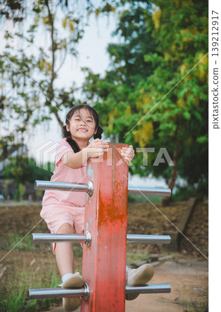 Happy child playing on outdoor climbing equipment with a joyful smile in a natural park during a sunny day surrounded by greenery and trees Happy child playing on outdoor climbing equipment with a joyful smile in a natural park during a sunny day surrounded by greenery and trees 139212917
