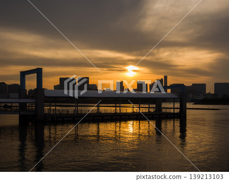 橘色的夕陽倒映在碼頭的水面上,遠處建築物的輪廓清晰可見(東京港區台場)。 橘色的夕陽倒映在碼頭的水面上,遠處建築物的輪廓清晰可見(東京港區台場)。 139213103