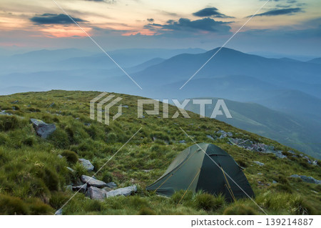 Summer camping in mountains at dawn. Tourist tent on round grassy hill on distant misty blue mountains range under pink sky before sunrise or sunset. Tourism, hiking and beauty of nature concept. 139214887