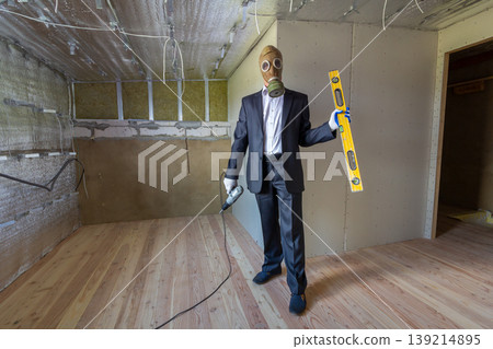 Strange man in businessman suit and gas protection mask inside a room under renovation works holding electric screwdriver and a level tools. 139214895