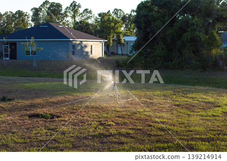 Sprinkler irrigating grass lawn with water in summer garden. Watering green vegetation during dry season for maintaining it fresh Sprinkler irrigating grass lawn with water in summer garden. Watering green vegetation during dry season for maintaining it fresh 139214914