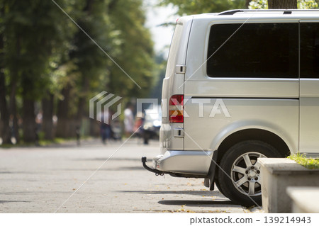 Side view detail of white passenger medium size luxury minibus van parked on summer city street pavement with blurred silhouettes of pedestrians on green trees bokeh copy space background. Side view detail of white passenger medium size luxury minibus van parked on summer city street pavement with blurred silhouettes of pedestrians on green trees bokeh copy space background. 139214943
