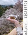 一個人手持櫻花餅（櫻花年糕），背景是盛開的櫻花和山景。 139215791