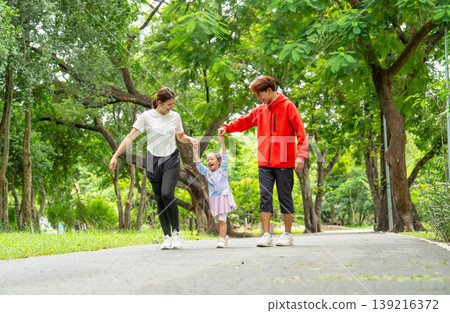 Front view Asian father, mother and daughter hold hands and walk together along the road in public park with day light, daughter look enjoy and fun with her family. Front view Asian father, mother and daughter hold hands and walk together along the road in public park with day light, daughter look enjoy and fun with her family. 139216372