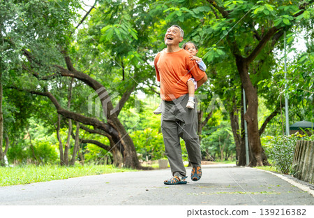 Asian grandfather carry the little girl with ride on his back and walk along the road in public park or garden and enjoy fun activity together with happiness. Asian grandfather carry the little girl with ride on his back and walk along the road in public park or garden and enjoy fun activity together with happiness. 139216382