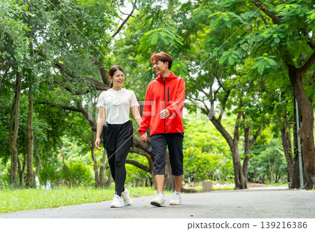 Asian man and woman hold hands and walk together along the road in public park with day light and they look happy with smiling. Asian man and woman hold hands and walk together along the road in public park with day light and they look happy with smiling. 139216386