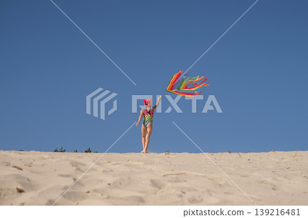A girl in a bright swimsuit runs along a sandy slope by the sea, launching an airy rainbow kite. Children's outdoor games. A Kid on the background of a clear blue sky A girl in a bright swimsuit runs along a sandy slope by the sea, launching an airy rainbow kite. Children's outdoor games. A Kid on the background of a clear blue sky 139216481