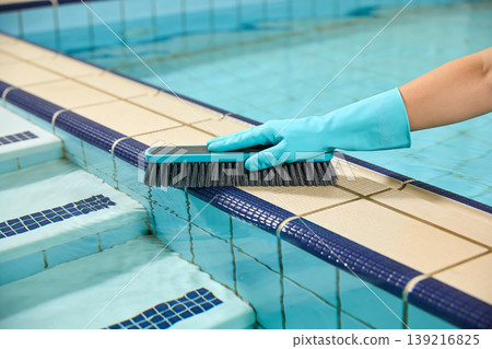 Hand in blue glove scrubbing pool edge tiles with a brush for cleaning and maintenance 139216825