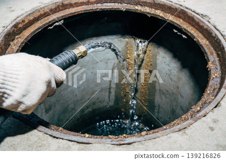 Worker in glove sprays water into an open sewer manhole with a hose 139216826