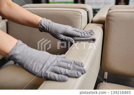 Close-up of hands wearing grey gloves cleaning a beige leather sofa armrest with a microfiber cloth 139216865